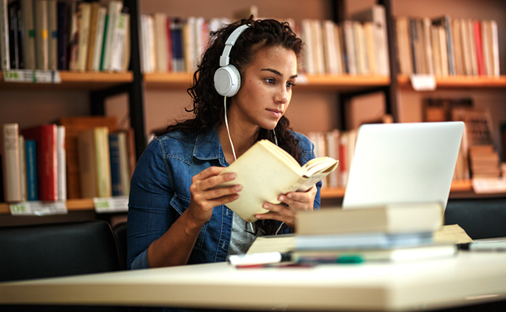 A young woman is holding a book and looking at a computer screen. She is wearing headphones.